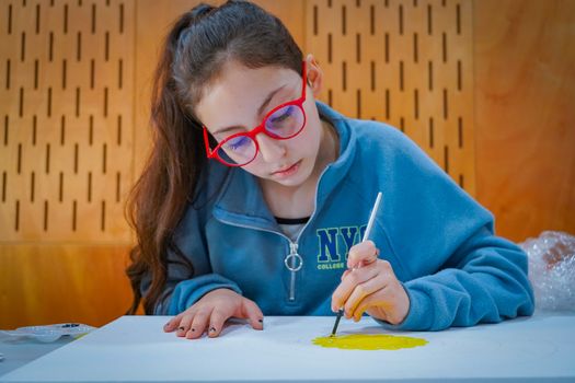 A young girl focused on painting while wearing bright red eyeglasses indoors.