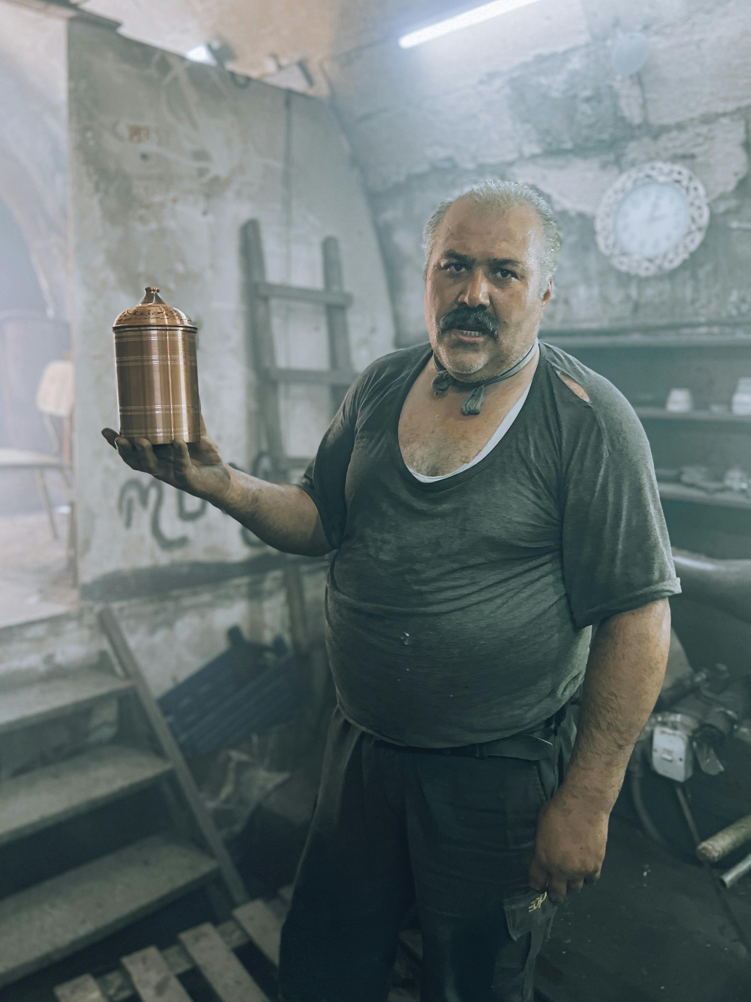 Middle-aged man holding a decorative container in a dusty, rustic workshop setting.
