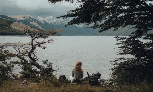 A tranquil scene of a woman sitting by a mountain lake, surrounded by nature.