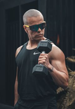 Muscular man in sunglasses lifts a dumbbell outdoors, showcasing strength and fitness.