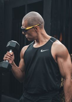 A muscular man performs a dumbbell workout outdoors in a tank top and sunglasses.