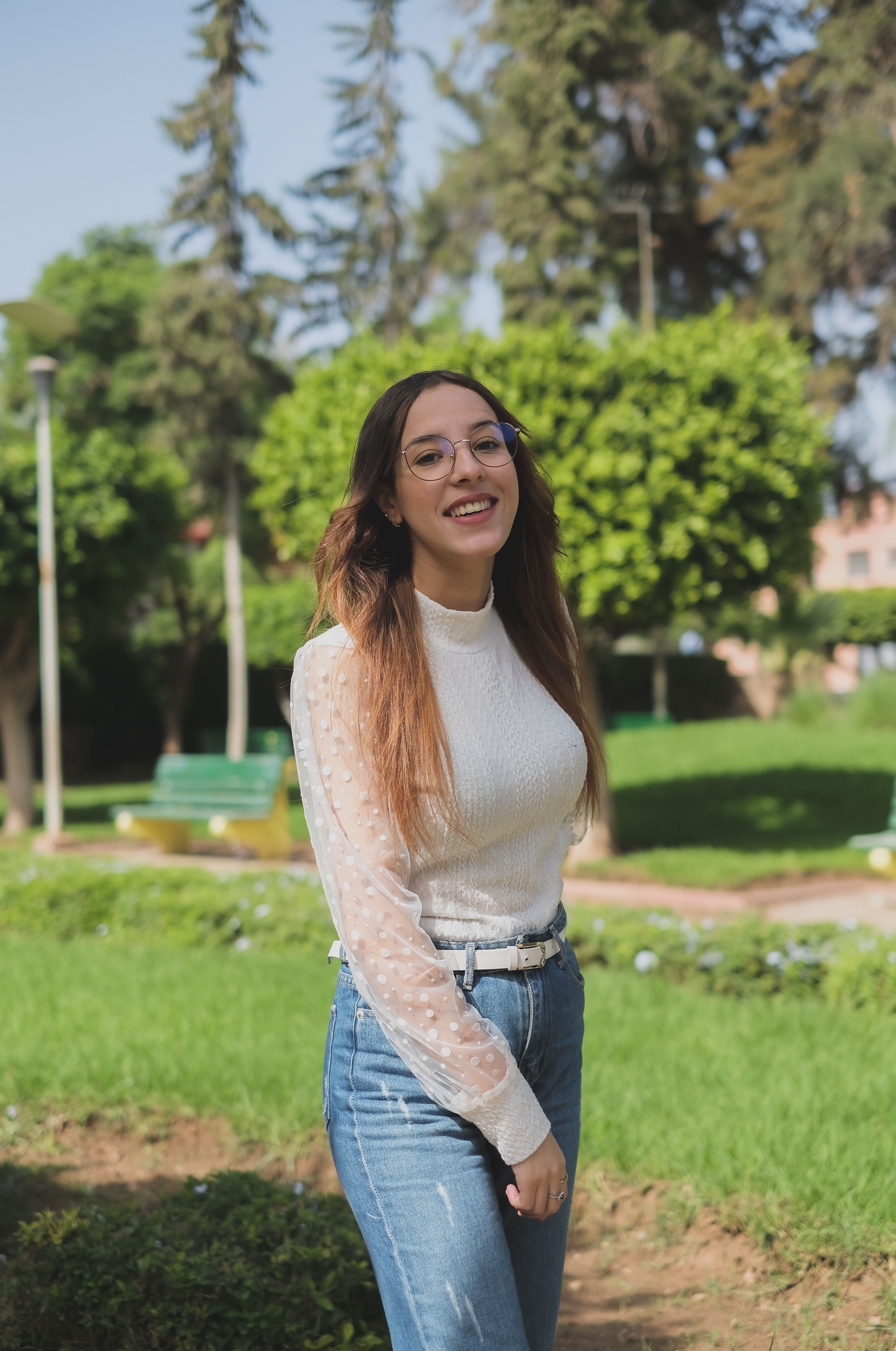 A young woman with eyeglasses smiles cheerfully in a sunlit park in Morocco.