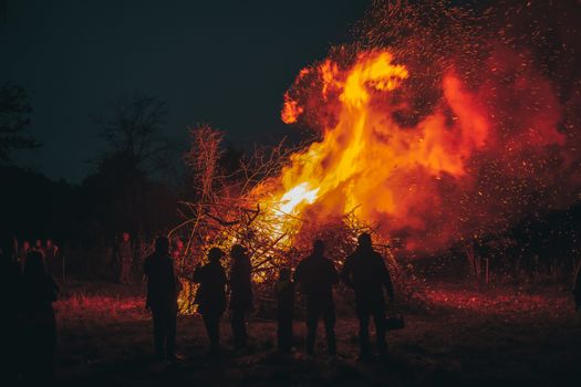 A blazing bonfire illuminates the night with silhouettes of people standing nearby.