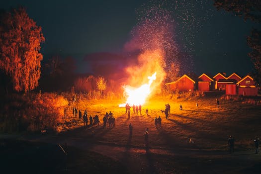 A large group enjoys a bright bonfire at night in a rural village setting, creating a warm and communal atmosphere.