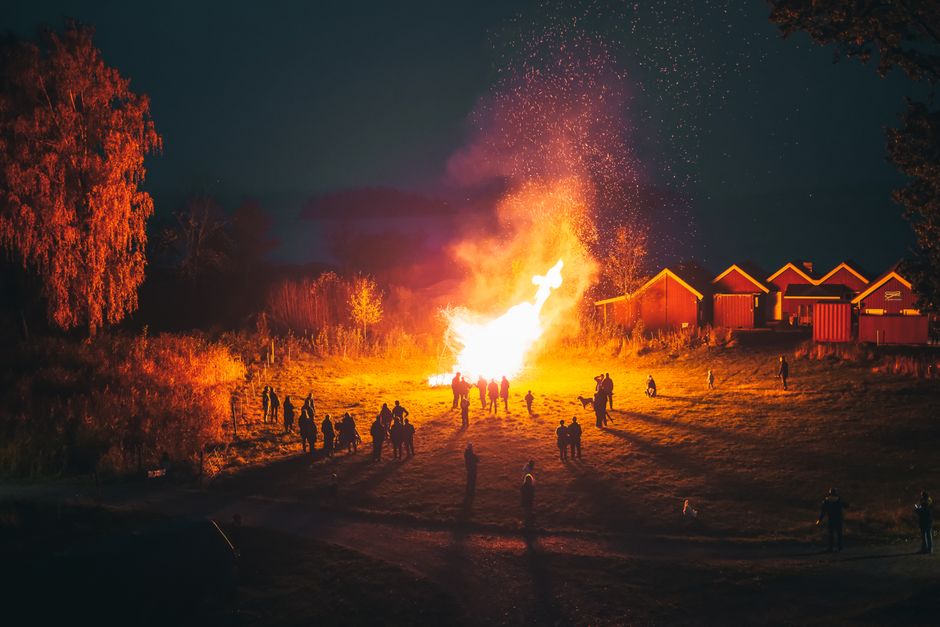 A large group enjoys a bright bonfire at night in a rural village setting, creating a warm and communal atmosphere.