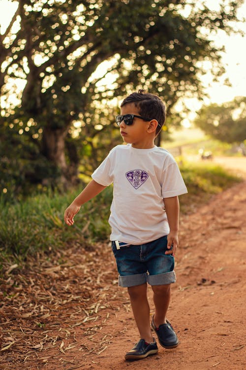 Boy in White Crew Neck T-shirt and Blue Denim Shorts Walking on Dirt Path