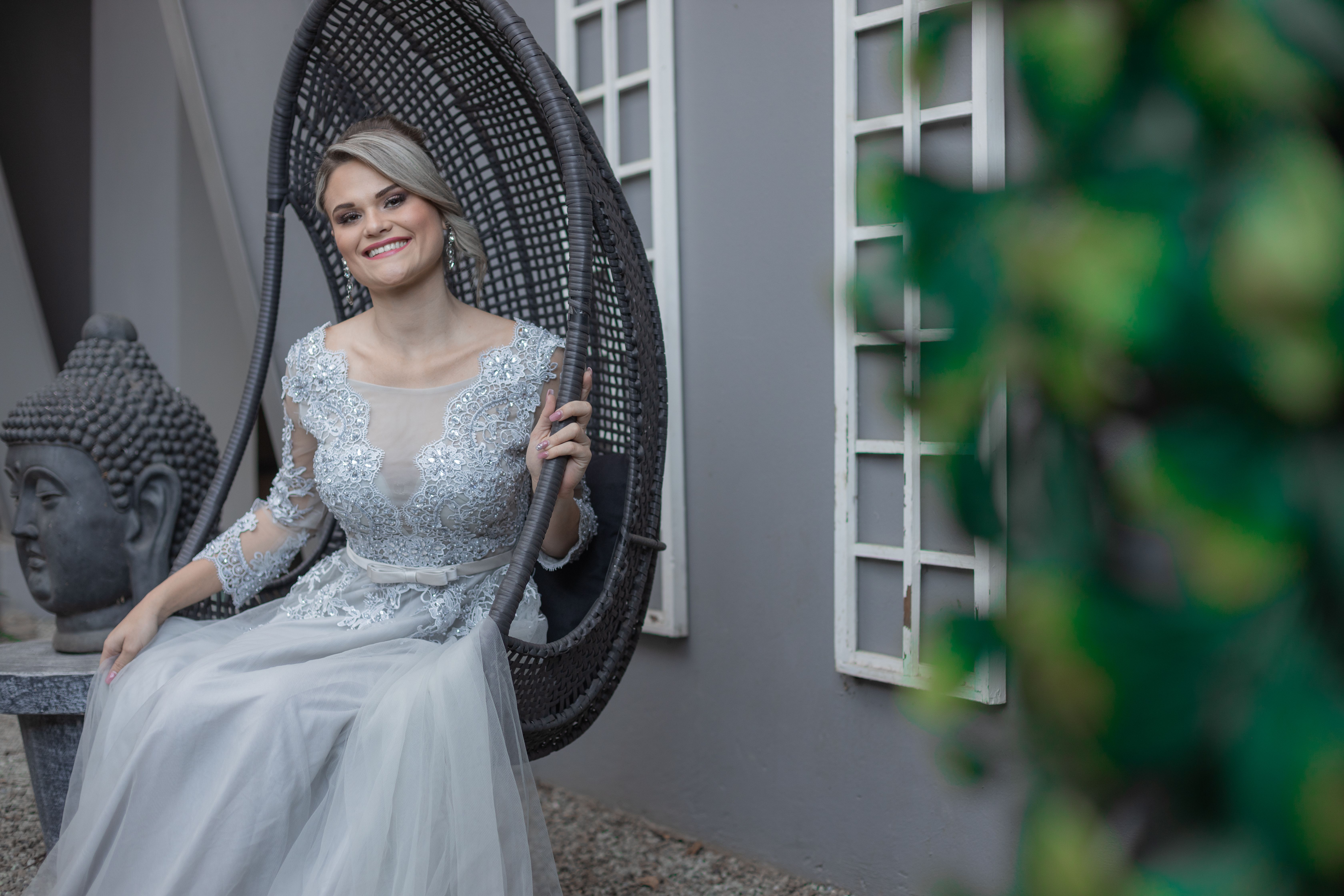 A smiling bride in a detailed lace wedding dress sitting in a hanging chair outdoors.