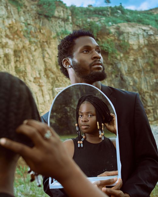 An artistic portrait of a man holding a mirror reflecting a woman, set against a rugged outdoor background.