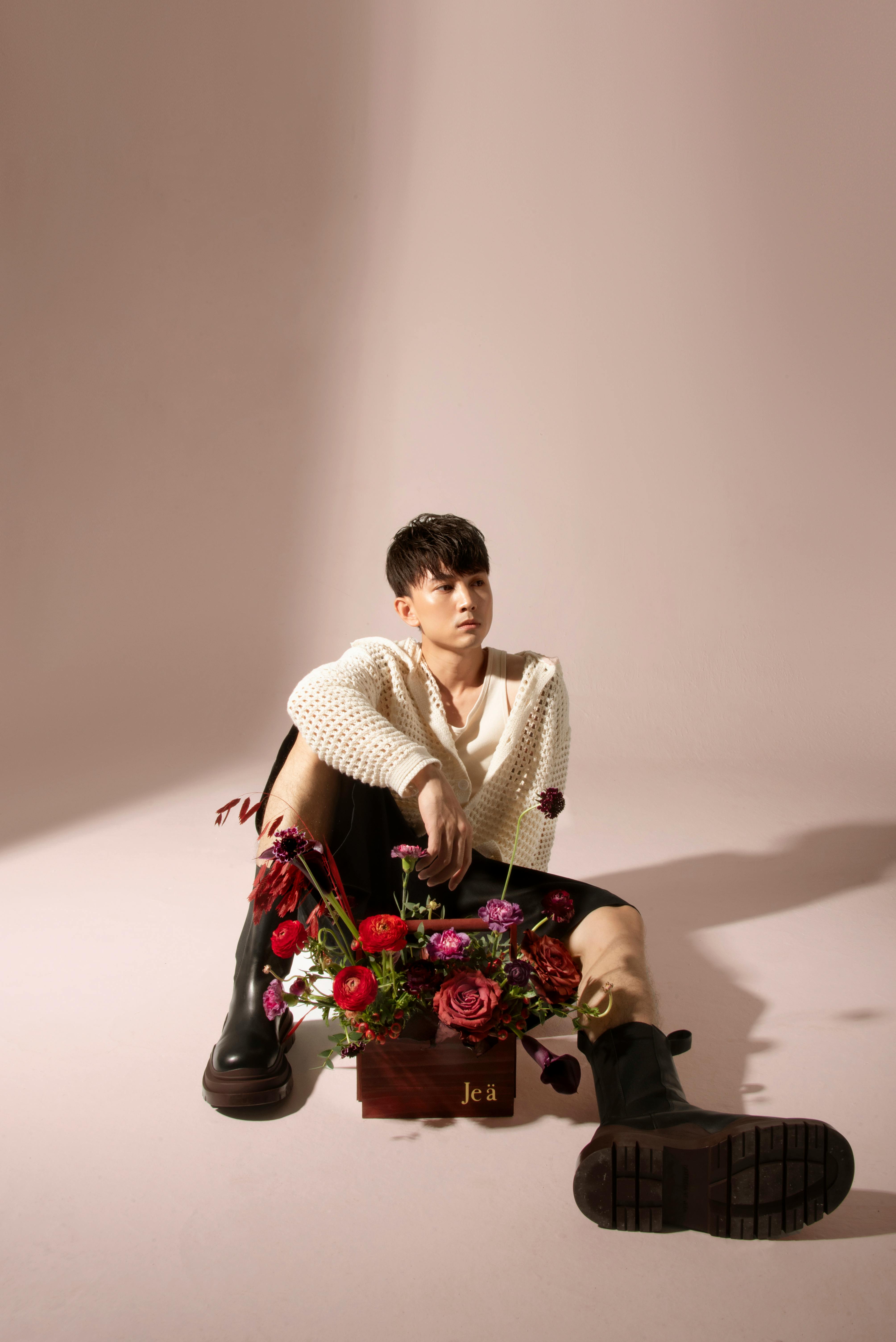 Man sitting in studio with bouquet, wearing stylish outfit and boots, posing creatively.