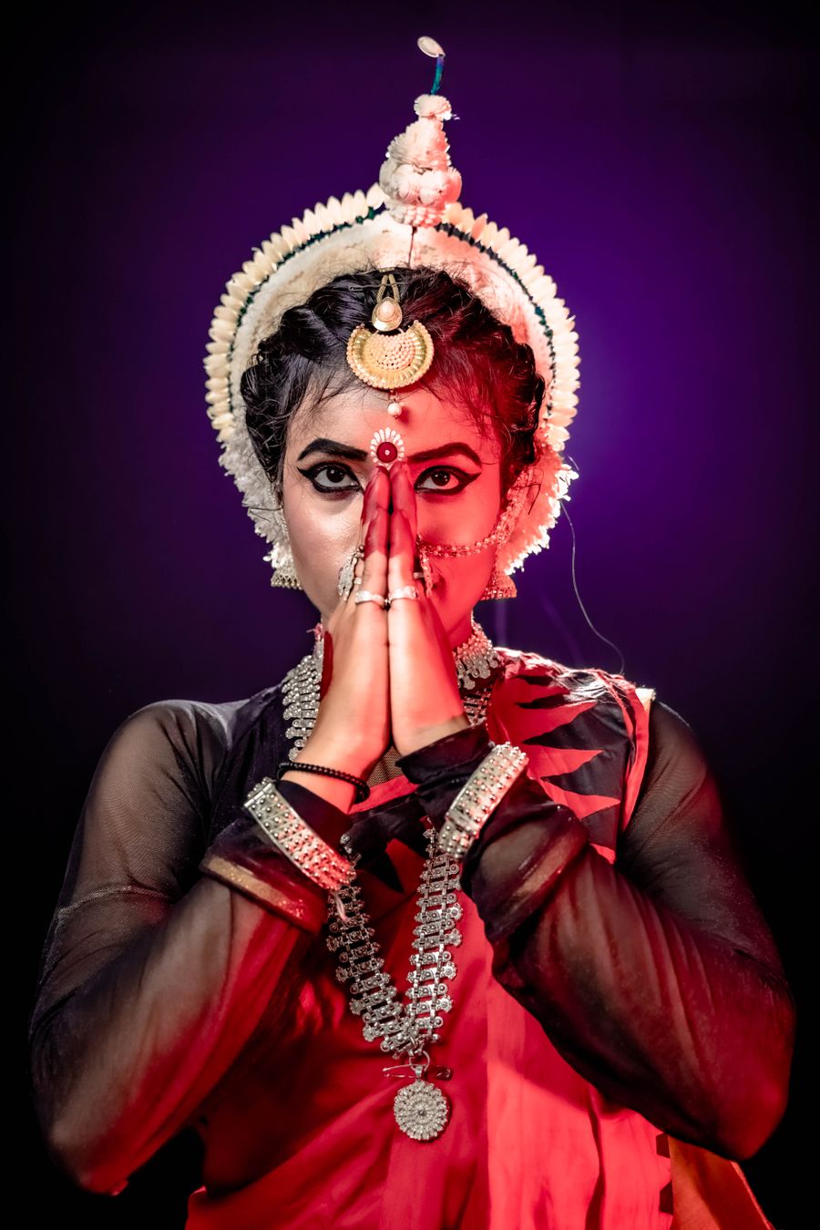 An Odissi dancer in full costume and silver headdress, hands joined in namaskar.