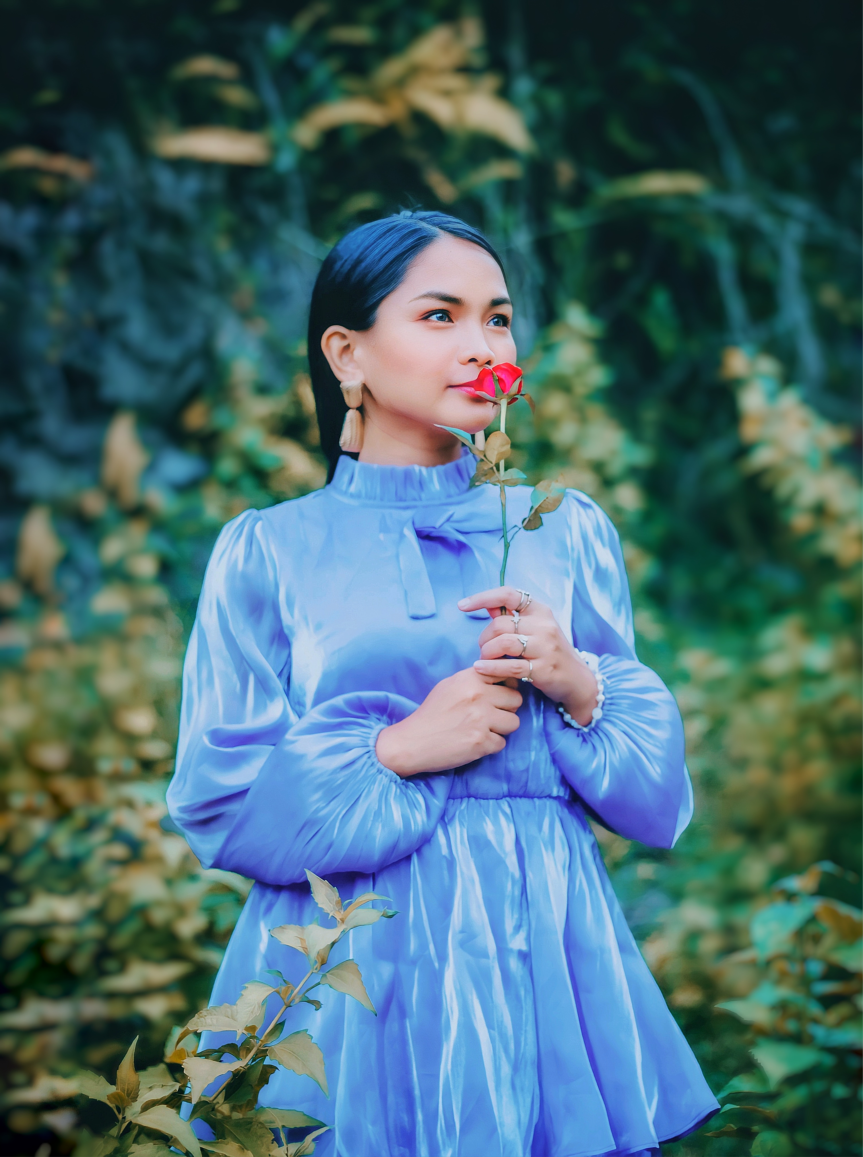 A woman in a blue dress holds a red rose in a lush outdoor setting, evoking elegance and tranquility.