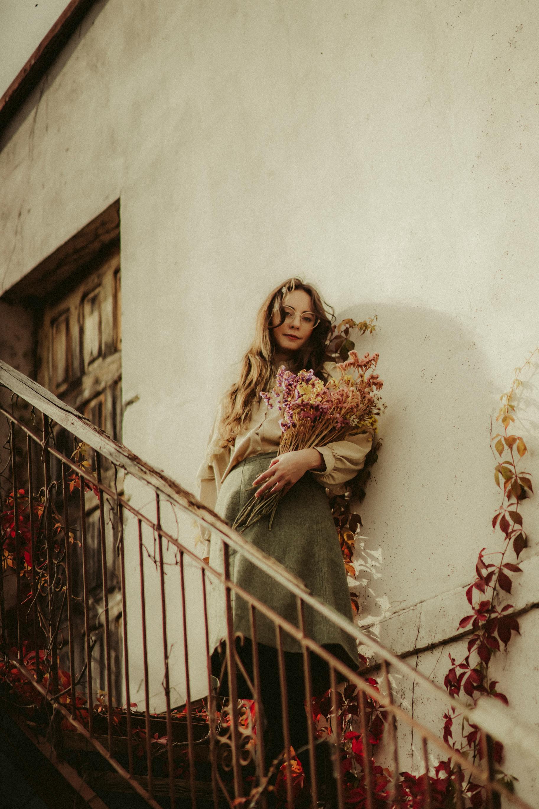 A woman stands on a rustic staircase holding a bouquet of flowers, surrounded by autumn foliage.