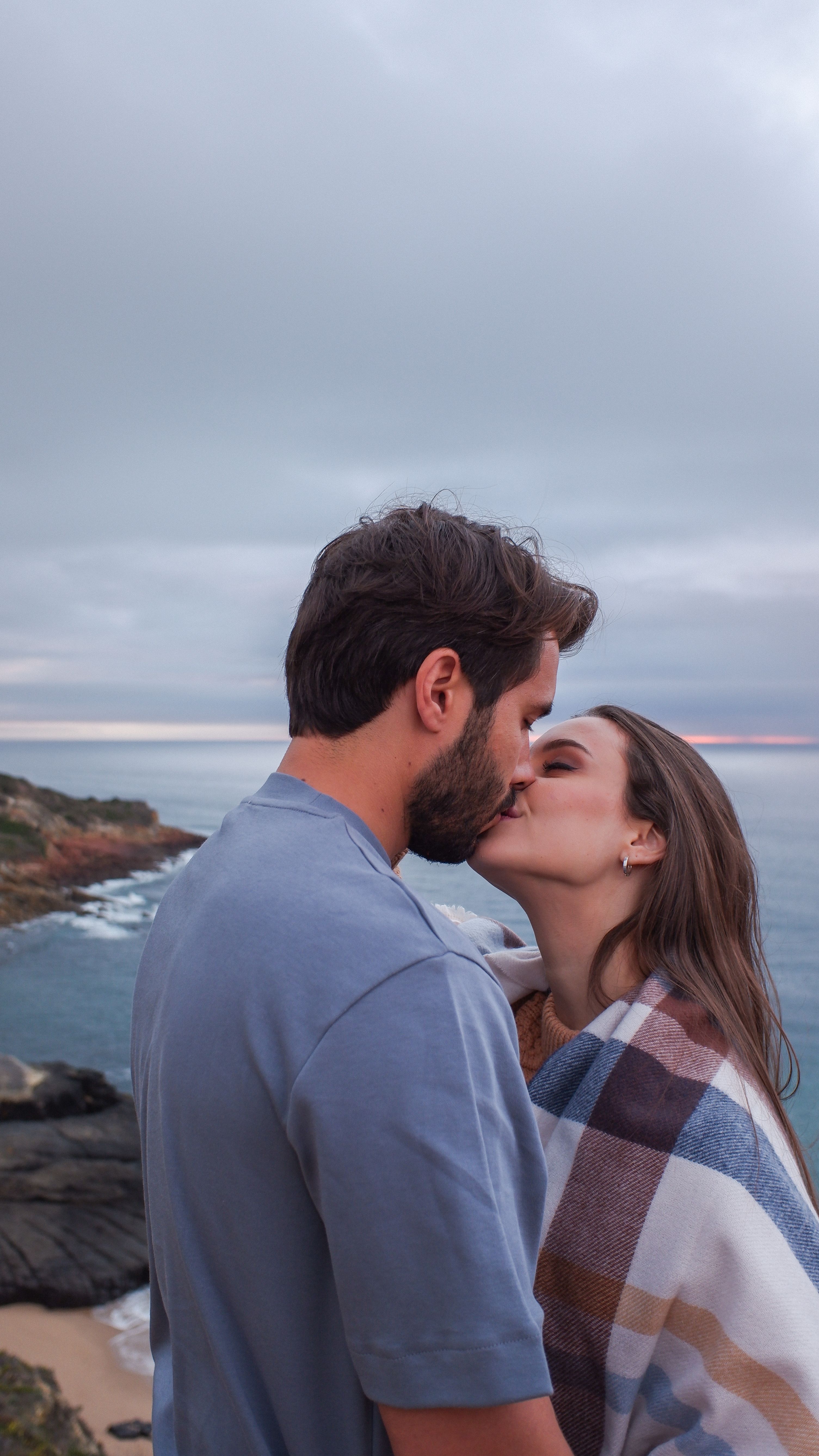 A couple shares a romantic kiss by the sea under a stunning sunset sky.