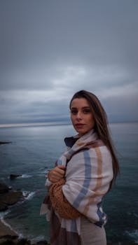 A woman stands wrapped in a blanket, gazing at the ocean during twilight. A serene and contemplative moment by the sea.