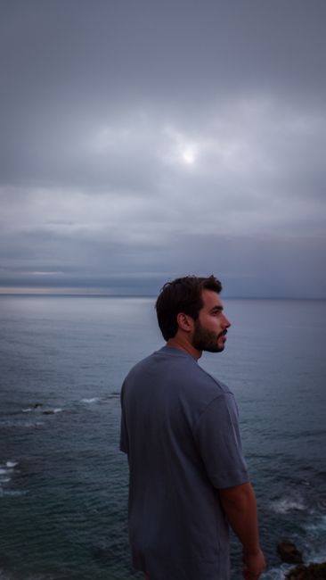 A contemplative man in a gray shirt overlooks the sea under a cloudy sky during twilight.