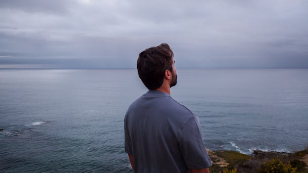A man stands on a cliff gazing at the peaceful ocean under a cloudy sky.