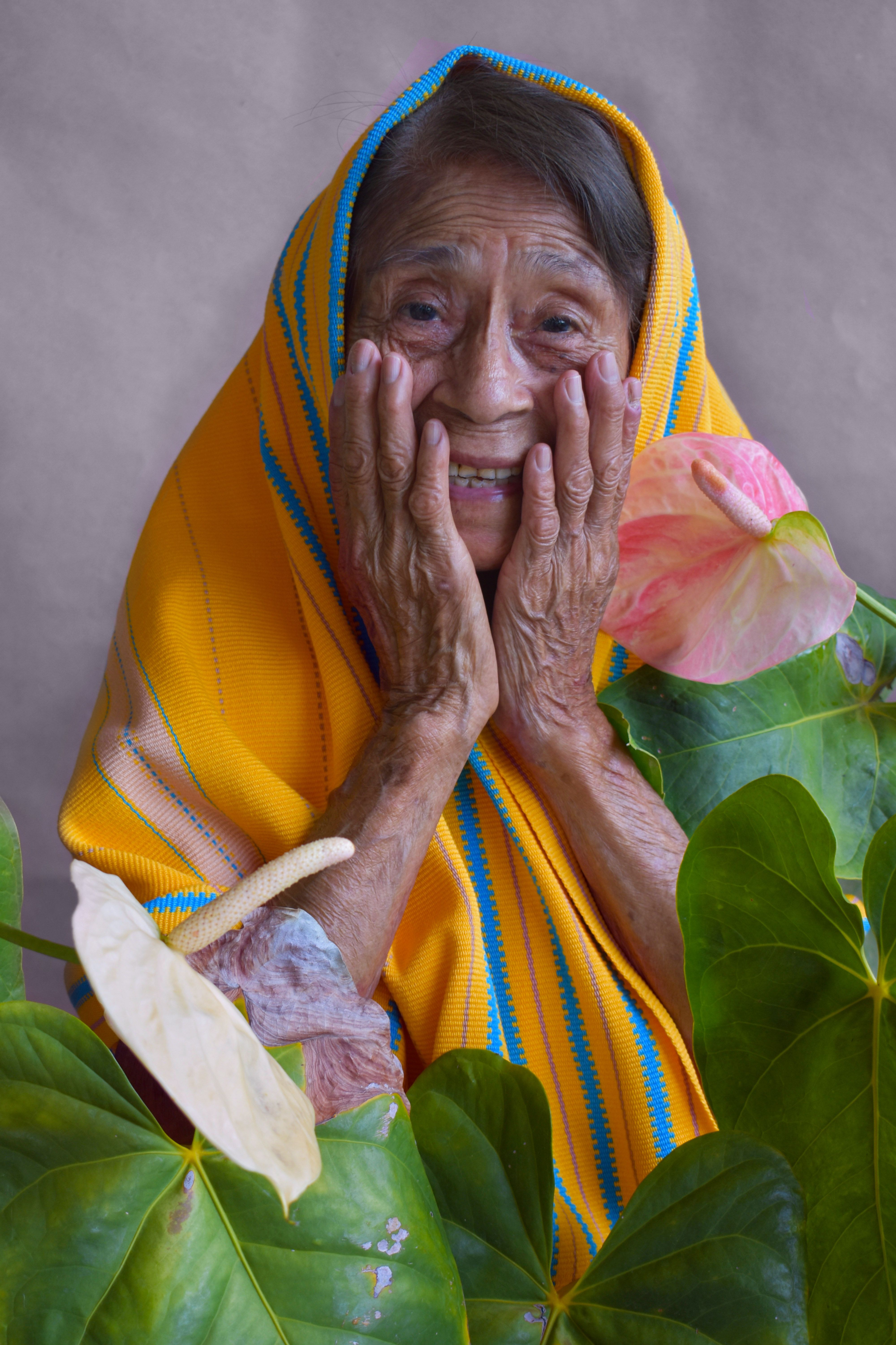 Charming elderly woman in vibrant headscarf surrounded by lush green plants, radiating warmth and happiness.