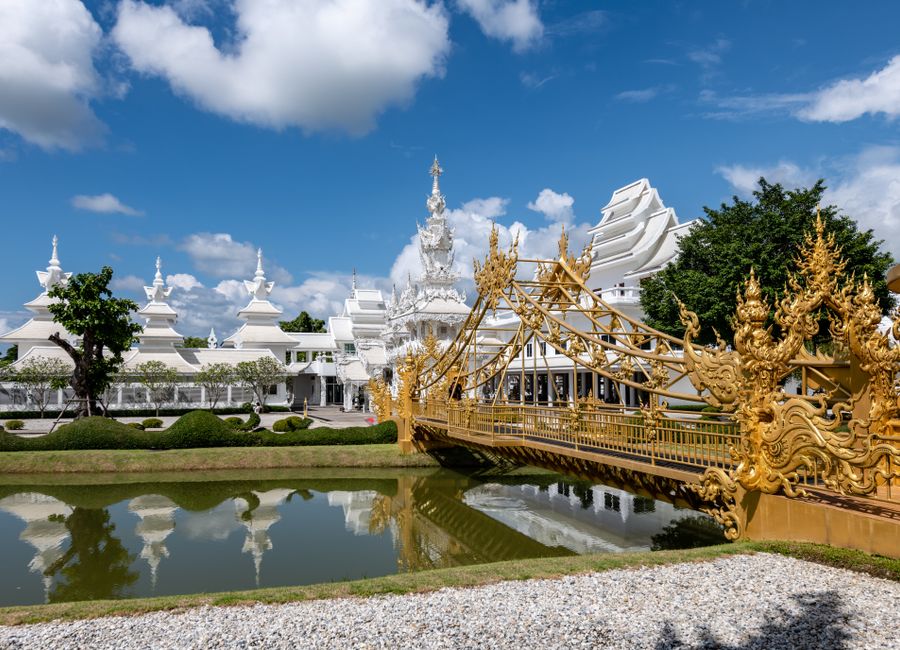 Golden bridge and Wat Rong Khun in Thailand, reflecting in a serene canal on a sunny day.