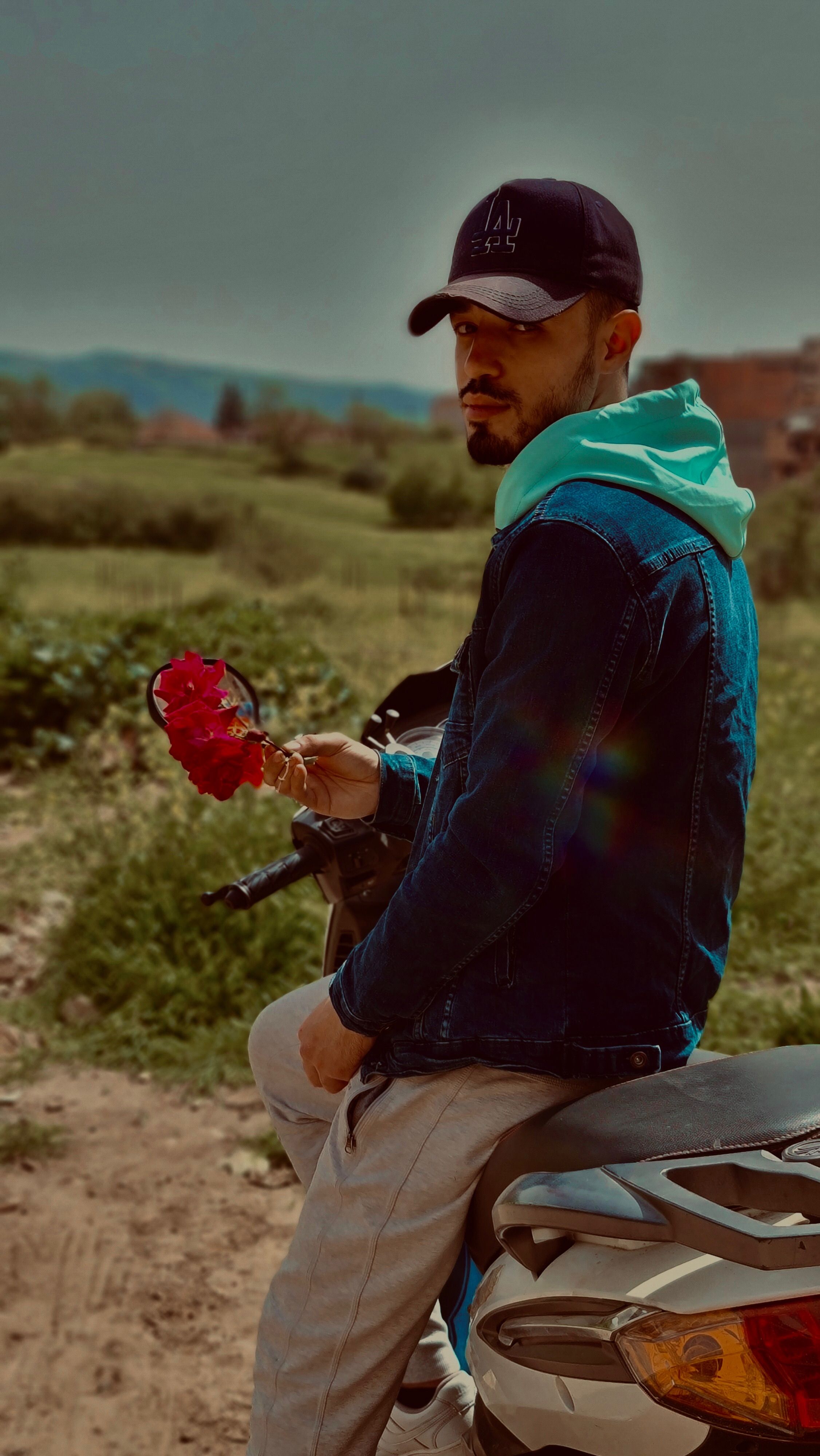 A man in casual clothing sits on a motorcycle, holding flowers in the Algerian countryside.