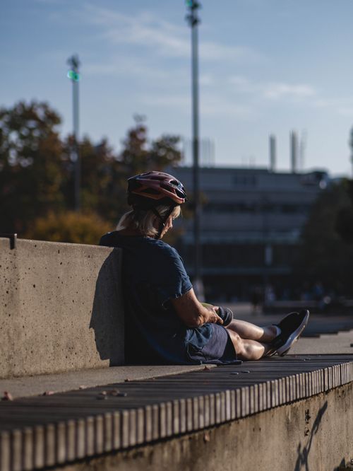 Free Elderly woman with bicycle helmet sits on a bench enjoying sunny urban scenery. Stock Photo