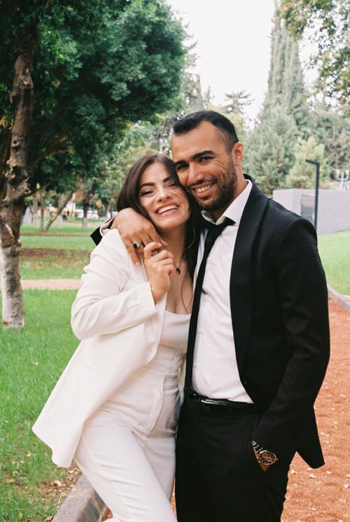 Free Happy couple in a park wearing formal suits, embracing and smiling under lush trees. Stock Photo