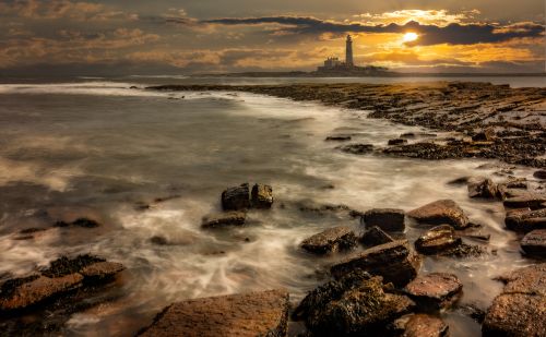 Free Dramatic sunset over rocky shore with distant lighthouse, waves creating a misty effect. Stock Photo