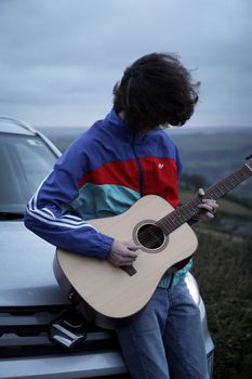 A young man plays guitar leaning on a car under a gloomy sky, creating a moody atmosphere.