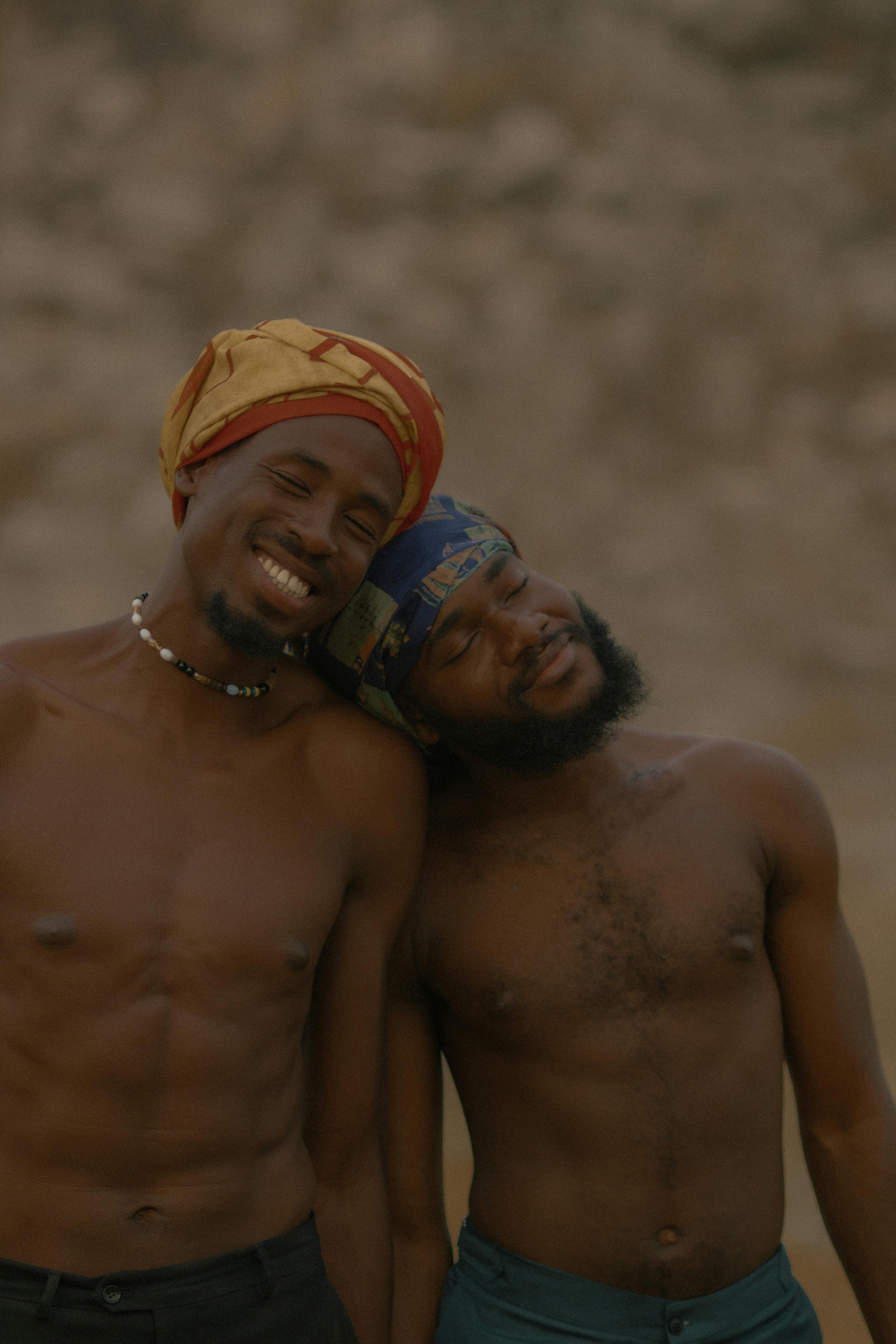 Two shirtless men with headscarves smiling and leaning on each other outside.