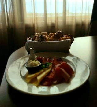 Plate of meats and cheese with bread basket by a window, warm morning setting.