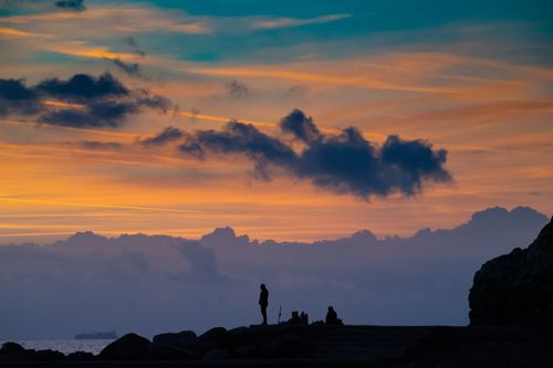 Free Silhouetted people on a rocky lakeshore under vibrant sunset skies with clouds. Stock Photo