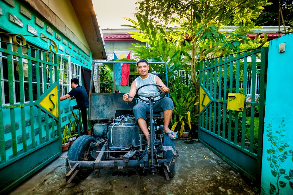 A man sits on a custom-built vehicle by a vibrant green gate with a Philippine flag.