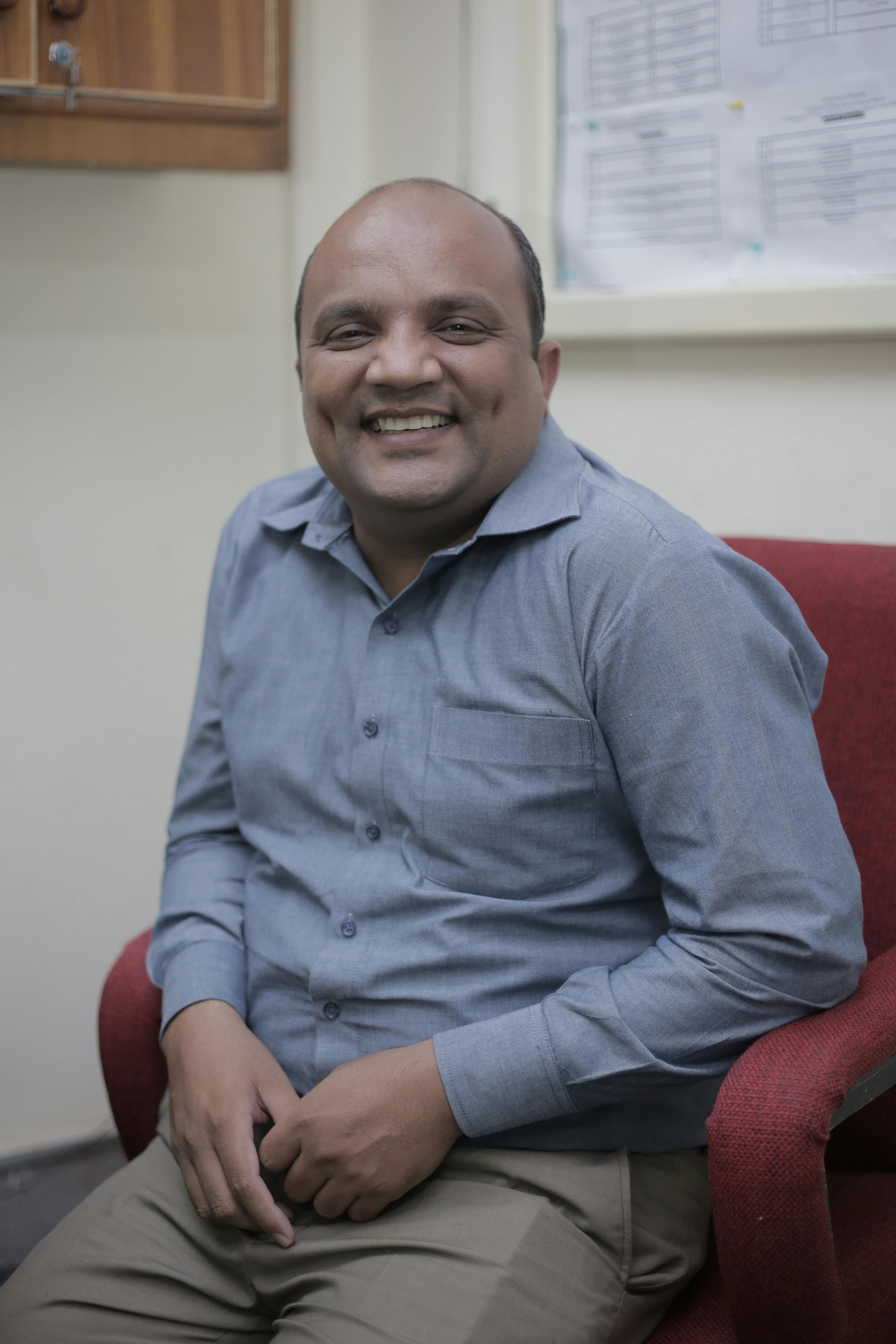 A smiling adult man in a blue shirt sitting on a red armchair indoors in Lahore, Pakistan.
