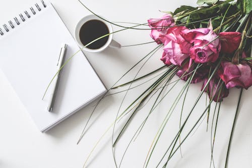 Ramo De Flores De Rosas Rojas Sobre Una Superficie Blanca Junto A Un Libro De Primavera Con Un Bolígrafo Y Una Taza De Cofffee