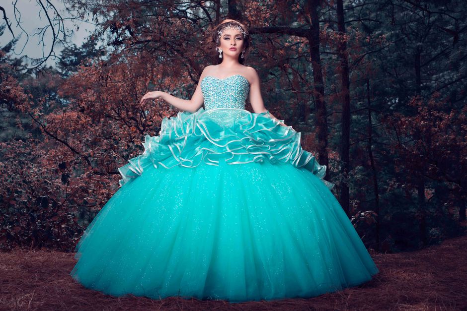 A woman in a glittering blue gown posing elegantly in a forest setting, showcasing fashion and grace.