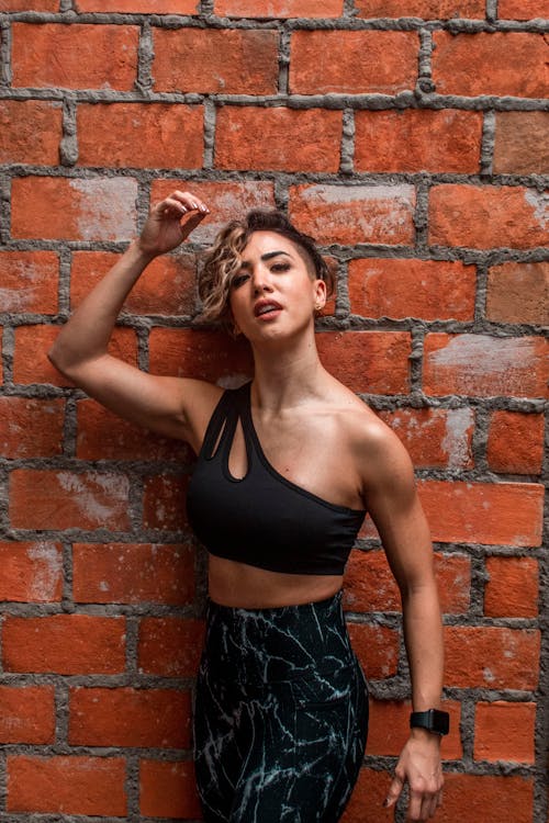 Woman in Black One Shoulder Sports Bra Standing Beside Brick Wall