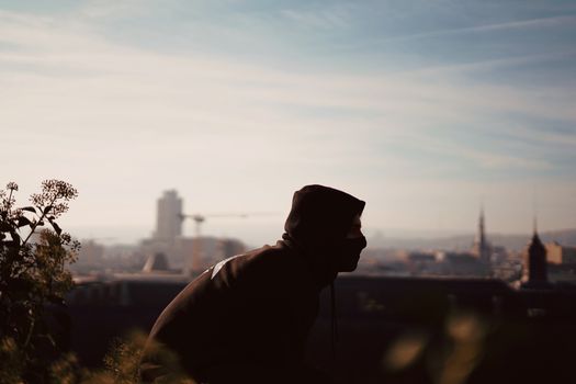 Silhouetted figure in hoodie overlooks urban cityscape at sunset, creating a moody atmosphere.