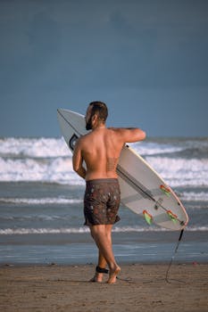 A man walks on the beach carrying a surfboard, ready for watersport action.