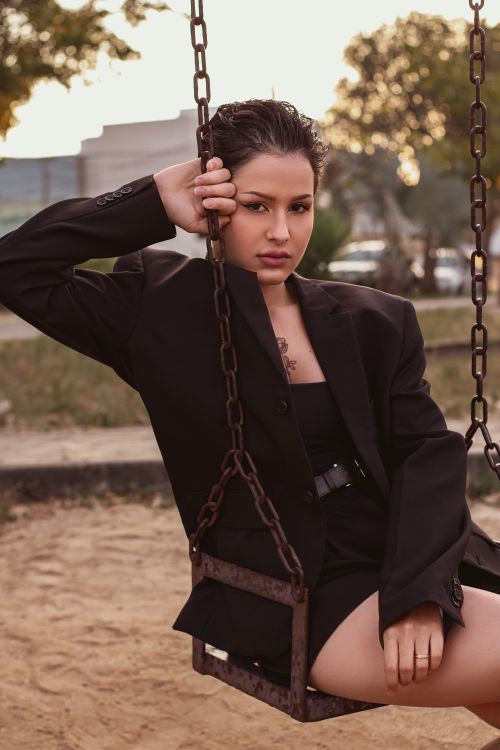 Free Confident woman in a black blazer posing on a swing in an outdoor setting. Stock Photo