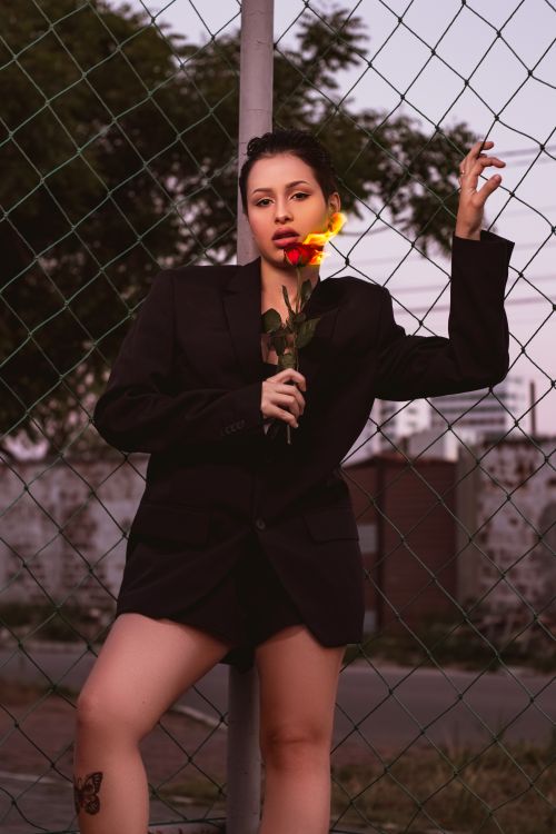 Free Young woman posing confidently with a red rose against a chain link fence backdrop. Stock Photo