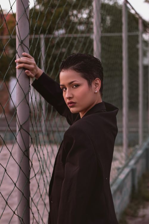 Free A teenager with short hair poses confidently by a chain link fence, exuding elegance and attitude. Stock Photo