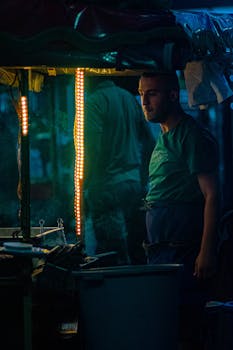 A vendor stands under illuminated lights at a night market with a thoughtful expression.