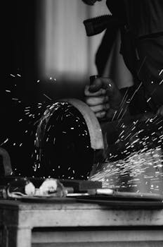 Black and white photograph of a craftsman creating sparks while cutting metal in a workshop.