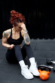 Redheaded woman in sportswear sitting in a gym, looking tired with tattoo and smartwatch visible.