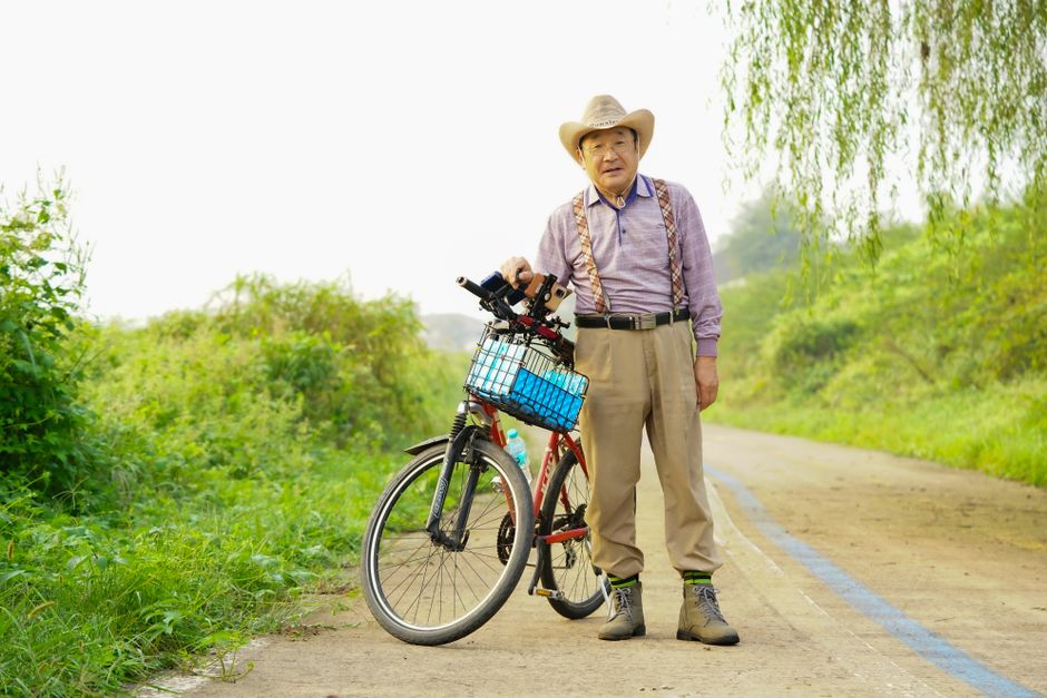 Elderly man standing with a bicycle on a rural path, wearing suspenders and a cowboy hat.