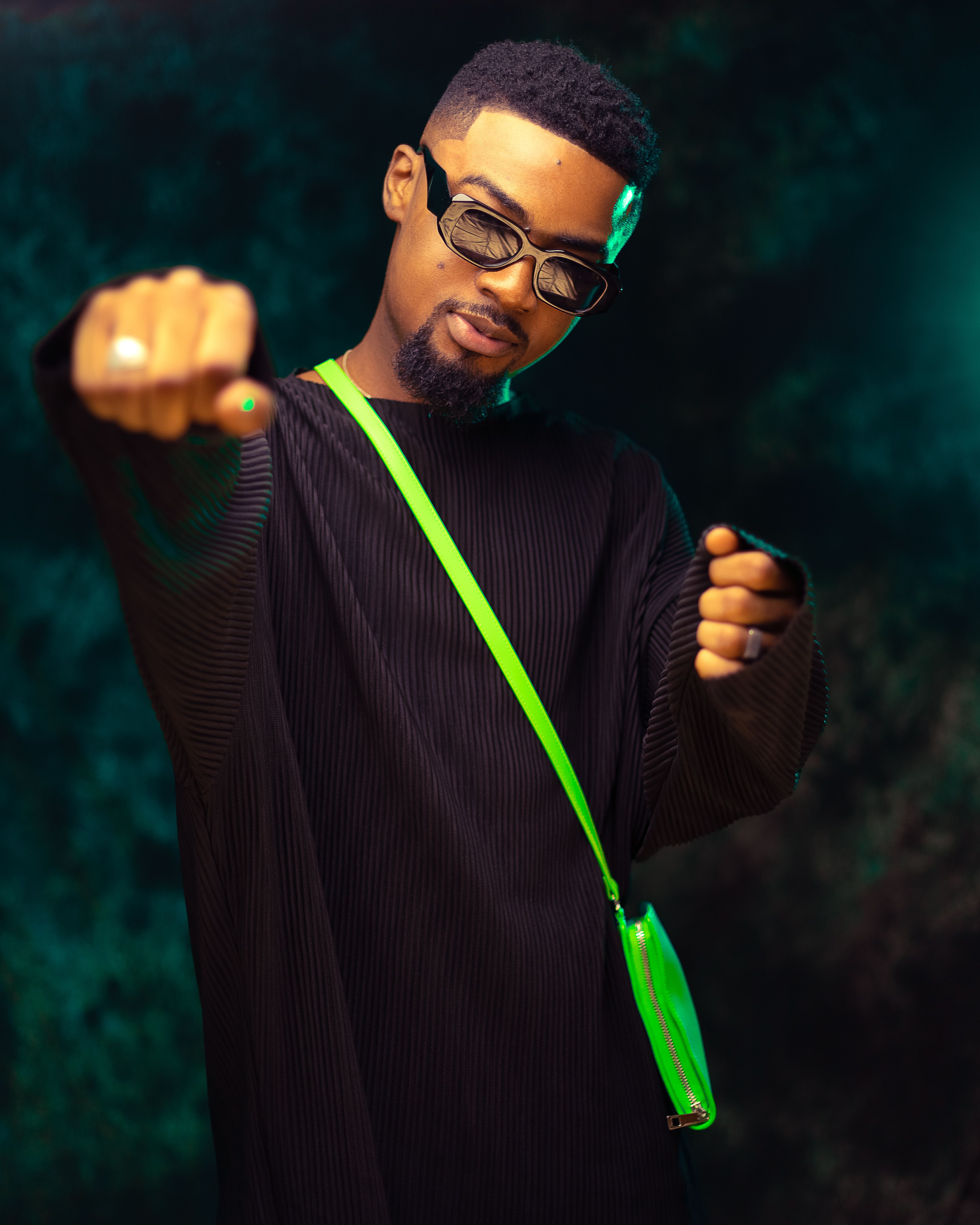 Fashionable portrait of a young man in sunglasses and a neon sling bag striking a confident pose indoors.