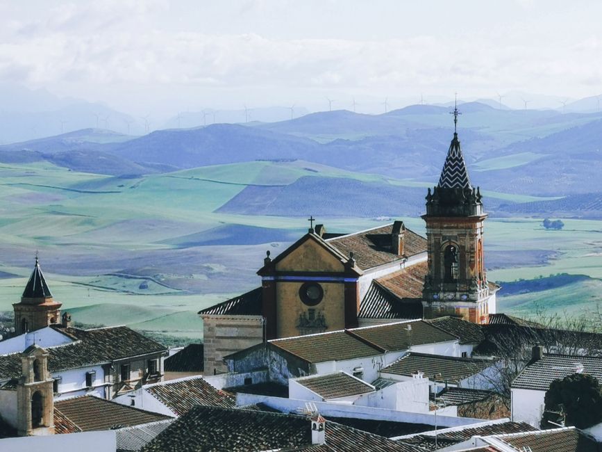 Una vista aérea escénica de una iglesia con colinas ondulantes y turbinas eólicas en un tranquilo paisaje de verano.