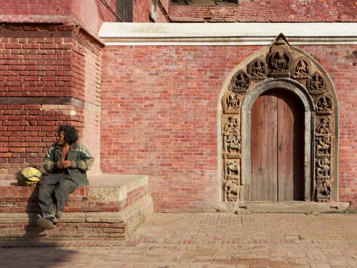 Free A man rests near an ornately carved red brick temple doorway, basking in the sunlight. Stock Photo