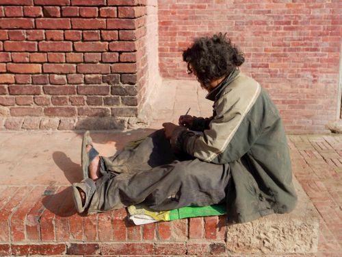 Free A homeless man with curly hair sits on a brick pavement writing with a pen. Stock Photo