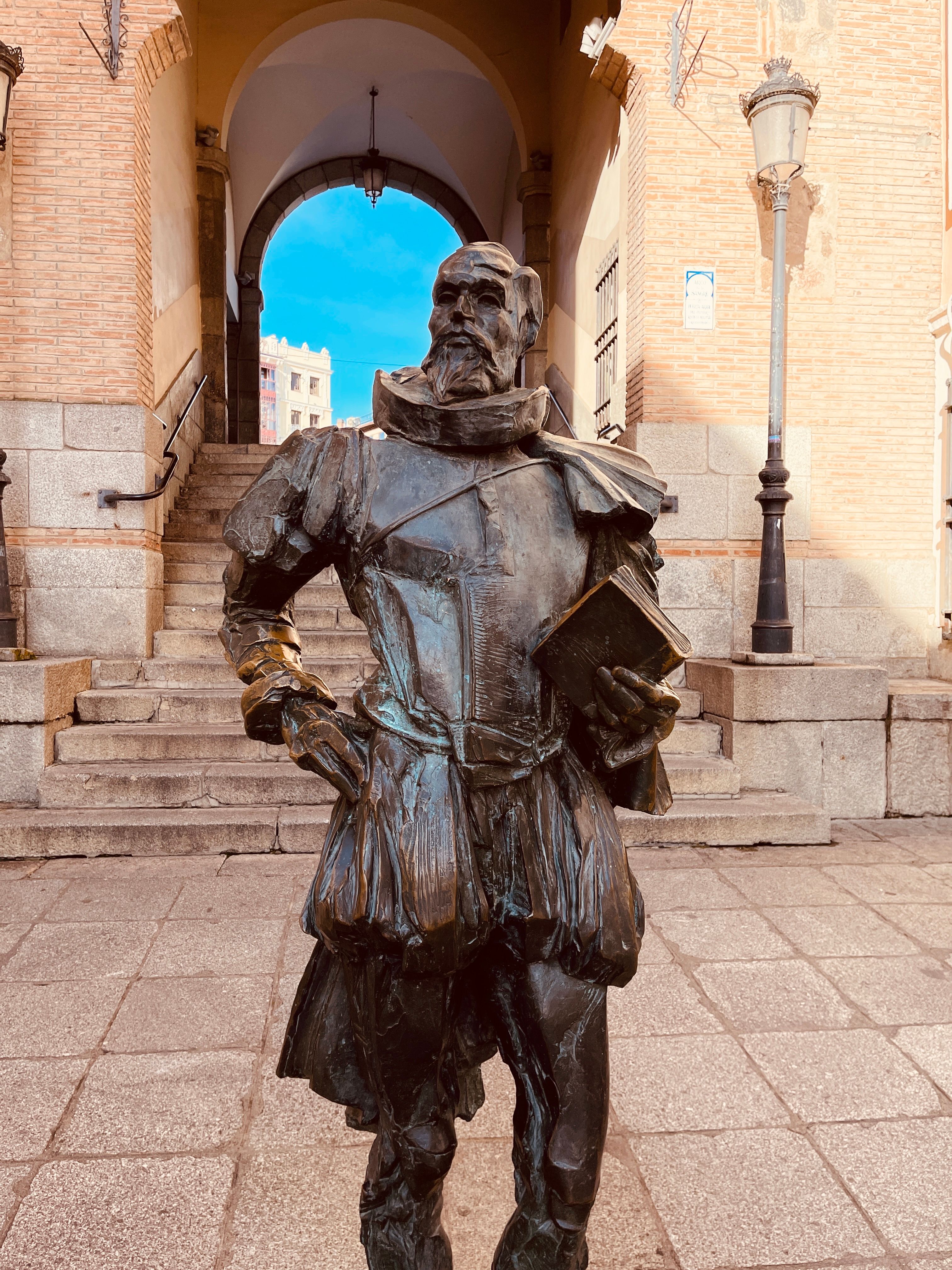 Bronze statue of a historical figure holding a book, set against a stone archway outdoors.