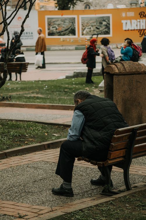 Free A man sits alone on a park bench, deep in thought as life bustles around him. Stock Photo